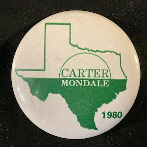 May include: A white button with a green outline of the state of Texas. The text "CARTER MONDALE" is printed in green inside the outline of the state. The year "1980" is printed in green at the bottom of the button.