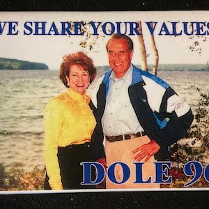 May include: A white button with a blue border and the text "WE SHARE YOUR VALUES" in blue letters. The button features a photo of a man and woman standing in front of a lake. The man is wearing a blue jacket over a white shirt and the woman is wearing a yellow blouse and black pants. The text "DOLE 96" is printed in blue letters at the bottom of the button.