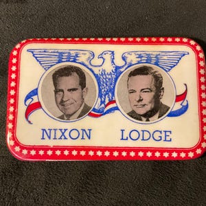 May include: A white button with a red border and white stars. The button features a blue eagle with outstretched wings and two portraits of men. The text "Nixon" and "Lodge" are printed below the portraits.