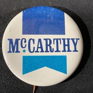 May include: A round, white button with the name "MCCARTHY" in blue lettering. The button has a blue rectangle at the top and a blue chevron shape at the bottom. A small blue star is next to the letter "C".