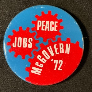 May include: A red and blue button with three interlocking gears. The gears have the words "JOBS", "PEACE", and "McGovern '72" on them.