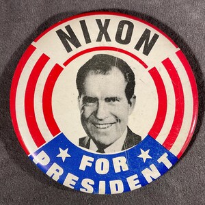 May include: A vintage political campaign button featuring a portrait of Richard Nixon. The button has a white background with red and white stripes, and blue text that reads "NIXON FOR PRESIDENT".