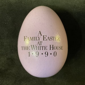 Puede incluir: Un huevo de color lila claro con el texto "A Family Easter at The White House 1990" impreso en negro. El huevo tiene una superficie lisa. El texto está centrado en el huevo, conmemorando un evento de Pascua en la Casa Blanca.