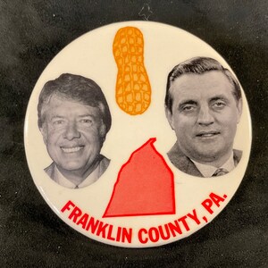 May include: A white button with a red outline. The button has a black and white image of two men, a peanut, and a red outline of a state. The text on the button reads 
