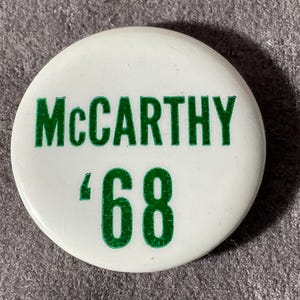 May include: White circular button with the name "McCarthy" in green block letters above the number "'68" in green. The button is a vintage political campaign button.