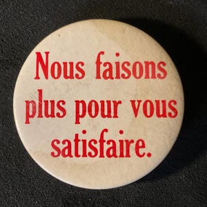 May include: A round, off-white button with the French text "Nous faisons plus pour vous satisfaire" in red lettering. The button has a slightly worn appearance, suggesting it is vintage or well-used.