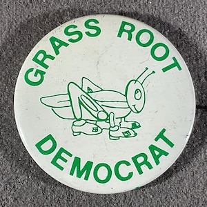 May include: A white button with green text and a green grasshopper illustration. The text around the edge reads "GRASS ROOT DEMOCRAT". The grasshopper is smiling and wearing shoes.