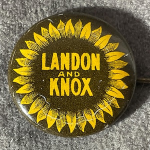 May include: A round pinback button featuring a dark brown background with a yellow sunflower graphic surrounding the text LANDON AND KNOX in bold yellow capital letters. The button has a metallic pin mechanism visible on the right side.