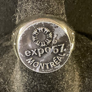 May include: Silver ring with a black circular design featuring the text "expo67 Montreal" and a stylized sunburst.