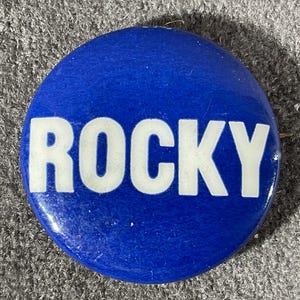 May include: A round, blue button with the white word "ROCKY" in bold, capital letters. The button has a pin on the back and is set against a textured, gray surface.