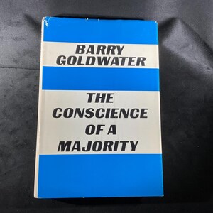 May include: A hardcover book titled "The Conscience of a Majority" by Barry Goldwater. The book cover features a white background with blue horizontal stripes and black text. The book is in good condition.