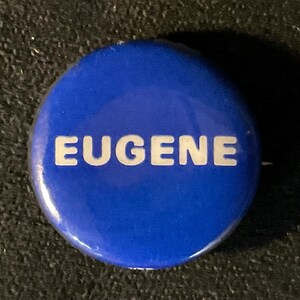May include: A round, blue button with the name "EUGENE" in white, capital letters. The button has a glossy finish and is set against a dark, textured background.