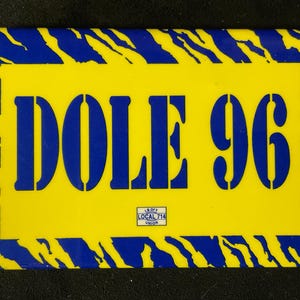May include: A rectangular yellow and blue sign with the text "DOLE 96" in blue block letters. The sign has a blue and yellow striped border. The text "LB OFF LOCAL 714 UNION" is in the lower center.