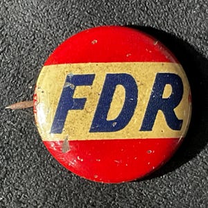May include: A circular pinback button featuring a horizontal yellow stripe across the center with the letters FDR printed in bold dark blue. The top and bottom sections of the button are red. The surface shows minor signs of wear and small scratches.