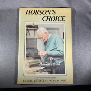May include: A book titled "Hobson's Choice" with an image of an older man working on a clock. The book cover is beige with black text. The man is wearing glasses and a green sweater. The text at the bottom reads "English Bracket Clock Repeating Work."