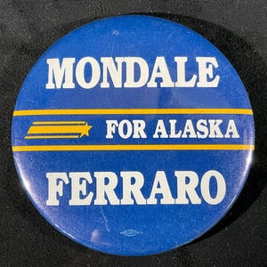 May include: A round, blue button with white text that reads "MONDALE FOR ALASKA FERRARO." The text is separated by a yellow horizontal stripe with a star and lines. The button is a vintage political campaign item.