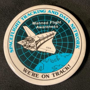 May include: A white button with a blue and black design. The button features a space shuttle, a radar screen, and the text "Spaceflight Tracking and Data Network, Manned Flight Awareness, We're on Track!"