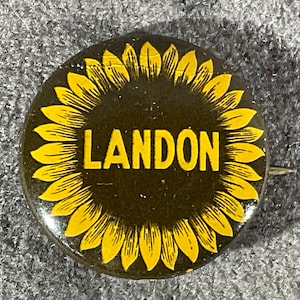 May include: A circular pinback button featuring a stylized yellow sunflower graphic against a dark brown background. The name LANDON is printed in bold, yellow, capital letters across the center of the sunflower design on the button face.
