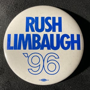 May include: A round, white button with blue text that reads "RUSH LIMBAUGH '96". The button has a simple, clean design with bold, sans-serif lettering. A small logo is at the bottom.