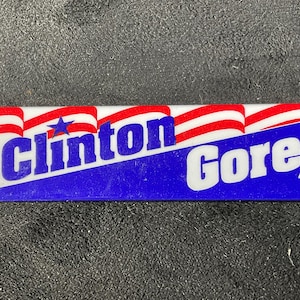 May include: Rectangular pin featuring the text "Clinton Gore '92" in white and blue. The design includes red and white striped flags and a blue star on a white background.
