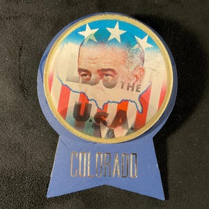 May include: A vintage campaign button featuring a portrait of a man, the text "THE USA," and the state name "COLORADO." The button has a blue ribbon shape with gold lettering and a circular image with red, white, and blue stripes.