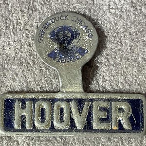 May include: Vintage metal pin with the word "HOOVER" in silver letters on a blue rectangular background. The pin has a circular top with the text "GREEN DUCK CHICAGO" and a blue graphic.