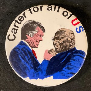May include: A white button with a blue and red border. The button features a photo of two men shaking hands. The text "Carter for all of us" is printed in black around the edge of the button.