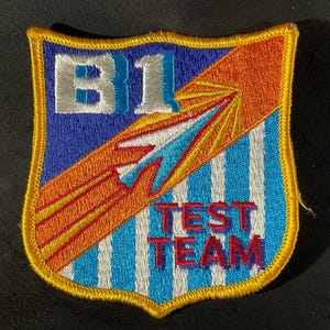 May include: Embroidered patch with a shield design. The patch features the text "B1 TEST TEAM" in white and red lettering. The shield is blue, orange, and white with a gold border. The design includes stripes and a stylized arrow.