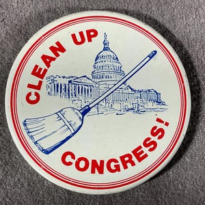 May include: Round white button with red and blue text and graphics. The button features the text "CLEAN UP CONGRESS!" around the edge, with a blue illustration of the U.S. Capitol building and a broom.
