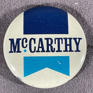 May include: A round button with a white background, featuring the name "MCCARTHY" in navy blue block letters. The button has a navy blue rectangle at the top and a light blue shape at the bottom. A small blue star is between the "M" and "C".