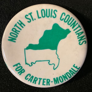 May include: A white, circular button with green text and a green outline of a geographical area. The text reads "NORTH ST. LOUIS COUNTIANS FOR CARTER-MONDALE". The button is a political campaign item.
