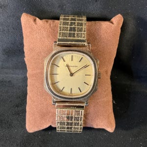 May include: A gold-toned wristwatch with a square face and a gold-toned metal band. The watch face has black Roman numeral hour markers and black hands. The watch face also has the word "Wittnauer" printed on it.