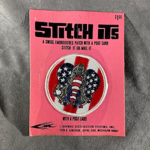 May include: A pink card with the text "STITCH ITS" and a Swiss embroidered patch featuring an elephant with American flag elements. The card reads "STITCH IT OR MAIL IT" and "WITH A POST CARD."