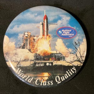 May include: A round button with a colorful image of a space shuttle launching. The button has the text "World Class Quality" and a NASA logo with the text "Manned Flight Awareness".