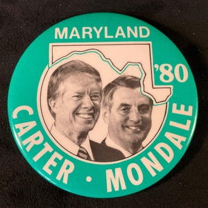 May include: A round, teal button featuring portraits of Jimmy Carter and Walter Mondale. The button includes the text "MARYLAND" at the top, the year "'80" and the names "CARTER" and "MONDALE" around the edge, with a white outline of the state of Maryland.