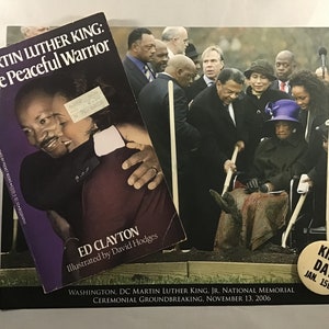 May include: A book titled "Martin Luther King: The Peaceful Warrior" by Ed Clayton, illustrated by David Hodges. The book cover features a photo of Martin Luther King Jr. embracing Coretta Scott King. The image also shows a photo of a groundbreaking ceremony for the Martin Luther King Jr. National Memorial in Washington, D.C. The ceremony took place on November 13, 2006.