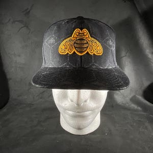 May include: A black baseball cap featuring a prominent embroidered golden-yellow bee emblem on the front panel. The fabric of the cap displays a subtle, repeating geometric honeycomb pattern across the crown and the flat brim.