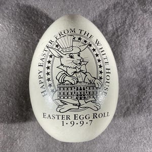 Puede incluir: Huevo color crema con una ilustración en blanco y negro de un conejo con sombrero de copa y pajarita, frente a la Casa Blanca. El texto dice "Happy Easter From The White House" y "Easter Egg Roll 1.9.9.7."