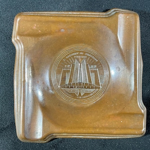 May include: A square, brown, glazed ceramic ashtray with rounded corners and four indented rests. The center features a circular embossed design depicting a stylized building with vertical lines and text reading A CENTURY OF PROGRESS CHICAGO.