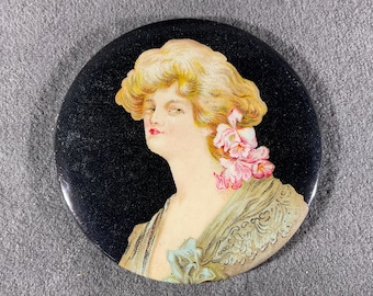 May include: Round black button with a vintage illustration of a woman with blonde hair and pink flowers. The woman is wearing a shawl with a light blue bow. The button has a glossy finish.