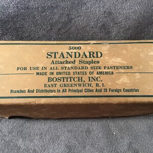 May include: A brown cardboard box with green text that reads "5000 STANDARD Attached Staples FOR USE IN ALL STANDARD SIZE FASTENERS MADE IN UNITED STATES OF AMERICA BOSTITCH, INC. EAST GREENWICH, R. I. Branches And Distributors in All Principal Cities And 29 Foreign Countries".