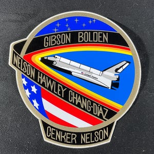 May include: A circular patch featuring a space shuttle design, with the words "GIBSON BOLDEN" at the top. The patch includes the names "NELSON HAWLEY CHANG-DIAZ" and "CENKER NELSON". The background has a blue sky with stars, a rainbow, and the American flag.