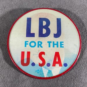 May include: A round button with a red border and the text "LBJ FOR THE U.S.A." in blue and red. The button has a white background with a faded image.