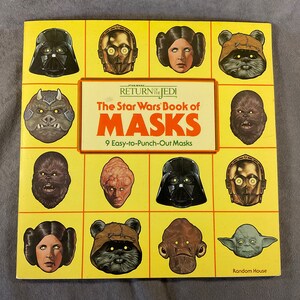 May include: A yellow book cover with the title "The Star Wars Book of Masks" and an image of nine Star Wars character masks, including Darth Vader, Princess Leia, Chewbacca, and Yoda. The book includes 9 easy-to-punch-out masks.