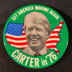 May include: A green campaign button with the text "GET AMERICA MOVING AGAIN" and "CARTER in '76". The button features a portrait of Jimmy Carter and a stylized American flag within a map outline.