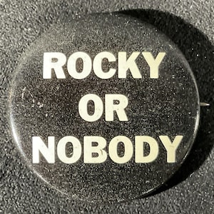 May include: A round, black button with the words "ROCKY OR NOBODY" in bold, white capital letters. The button has a pin on the back and is likely a collectible or promotional item.