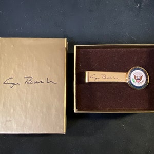 May include: A gold tie clip with the Presidential Seal and the signature "George Bush" on a gold background. The tie clip is in a gold box with the signature "George Bush" on the front.