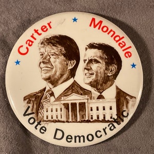 May include: A white button with a brown and white image of Jimmy Carter and Walter Mondale. The text "Carter Mondale" is in red letters and "Vote Democratic" is in black letters. There are two blue stars on the button.