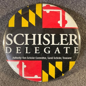 May include: A black and white button with the text "SCHISLER DELEGATE" in white. The button also features the Maryland state flag in the background.