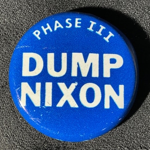 May include: A round, blue button with white text. The text reads "PHASE III" at the top, and "DUMP NIXON" in a larger font below. The button is set against a dark, textured background.